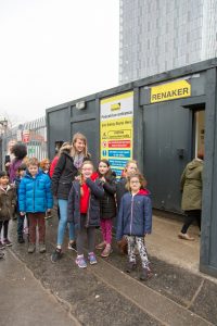 Photo of children on visit to Renaker construction site. They are standing in front of a black static caravan with a yellow 'Renaker' sign on it.