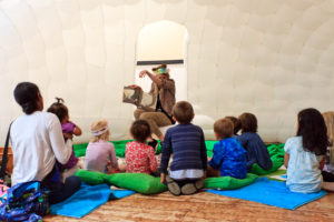 A woman dressed in animal print is sitting in front of a group of children reading a story to them. The children are sat on green cushions and blue mats.