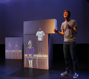 Production image from Friends For All. A man is standing in front of projected images of children who are wearing school uniform, one child is stood on a table wearing a white t-shirt that says 'fight the power' on it),