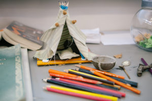 Picture of a small teepee tent made of push pins, beige fabric and pencils. The toy tent is behind a pile of coloured pencil crayons.