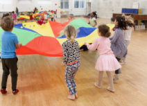 Lead image for Visiting Z-arts page. Picture of a group of 4 - 6 year olds standing in a circle holding a multi-coloured parachute. There are small multi-coloured balls bouncing on the parachute and the children are stood on a wooden, laminate floor.