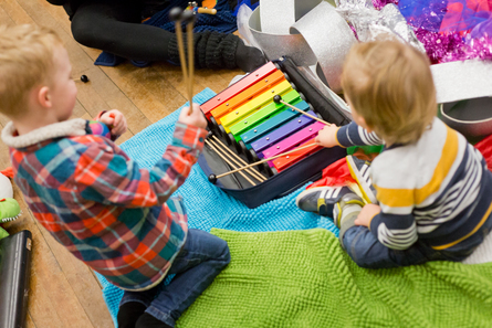 Two pre-school aged children are sat on green and blue mats playing on a rainbow coloured xylophone.