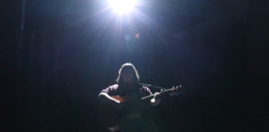 Photo of a woman holding a guitar. She is standing under a spotlight and silhouetted by the light.