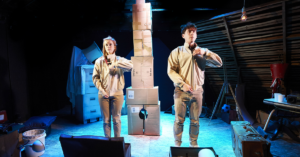 One Small Step production image. A man and a woman dressed in beige are facing forward, zipping up their jackets in unison. In the background is a tower made of cardboard boxes.