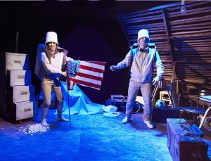 Production shot - One Small Step. A man and a woman dressed in beige are wearing buckets on their heads. The woman is holding an american flag and they are both wearing cardboard boxes as though they are rucksacks