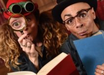 Picture of two people reading a book. One is holding a magnifying glass up to their eye and is wearing a pair of binoculars. The other is wearing a black bowler hat and wearing glasses.