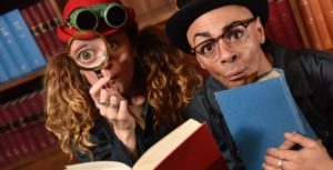 Picture of two people reading a book. One is holding a magnifying glass up to their eye and is wearing a pair of binoculars. The other is wearing a black bowler hat and wearing glasses.