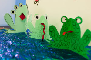 Picture of green paper frogs with red tongues and big eyes made by children and families as part of Family Art Stars. The frogs are arranged on a piece of shiny, sparkly, blue paper.