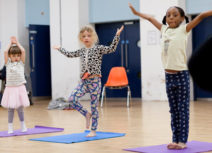Three young girls are standing on yoga mats and holding their arms up in the air.