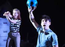One Small Step Production photo. In the foreground a man with short dark hair wearing a blue shit and black bow tie is holding up a black and blue football. Behind him a woman in a black and white striped tshirt is holding a toy spaceship.