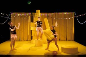 Production image from Tidy Up, three dancers wearing black tshirts and bronze shorts stand on a set with yellow curtains, fairy lights and large yellow sponge shapes.