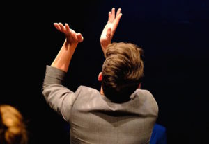 A man with short brown hair wearing a grey blazer is facing away from the camera and clapping into the air.