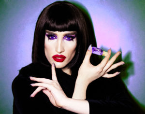 Picture of a woman with black hair, with purple eyes and red lipstick holding a small vial of purple liquid with a cork top.