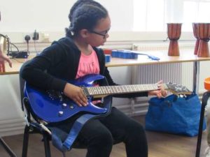 A girl is sat wearing a black cardigan, pink tshirt and black leggings playing a blue electric guitar.
