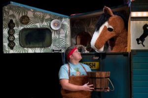 Production photo from Black Beauty. A man holding a bucket wearing a red headband and blue tshirt is looking up at a brown and white pantomime horse who is inside a blue horsebox.