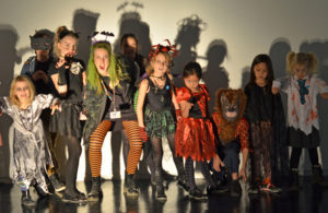 Children and an artist stand in a line in different halloween costumes making scary faces at the camera