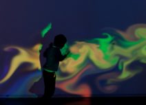Photo of a colourful, psychelic style projection of yellow, green, red and blue on a wall silhouetting a young child standing in front of it.
