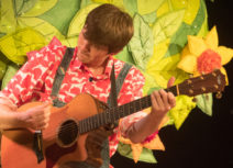Production Image - Climb That Tree. Man with short brown hair playing a guitar and wearing dungarees and a red and white patterned shirt.