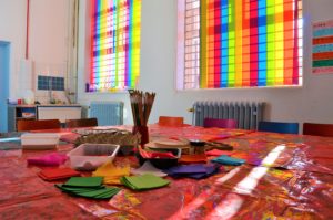Lead image for About Us page. Photo of Z-arts gallery workspace. A table is covered in paint-stained shiny red tablecloths with cardboard, felt, paintbrushes and other art resources on it. In the background there are rainbow coloured blinds covering two windows.