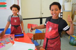 Two children are wearing red, paint-stained aprons and doing art activities. One of the children is looking directly at the camera and is doing a thumbs up.