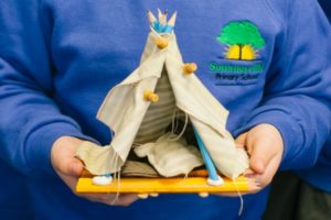 A child wearing a blue school sweatshirt is holding a small toy teepee-style tent made of pencils, push-pins and beige fabric