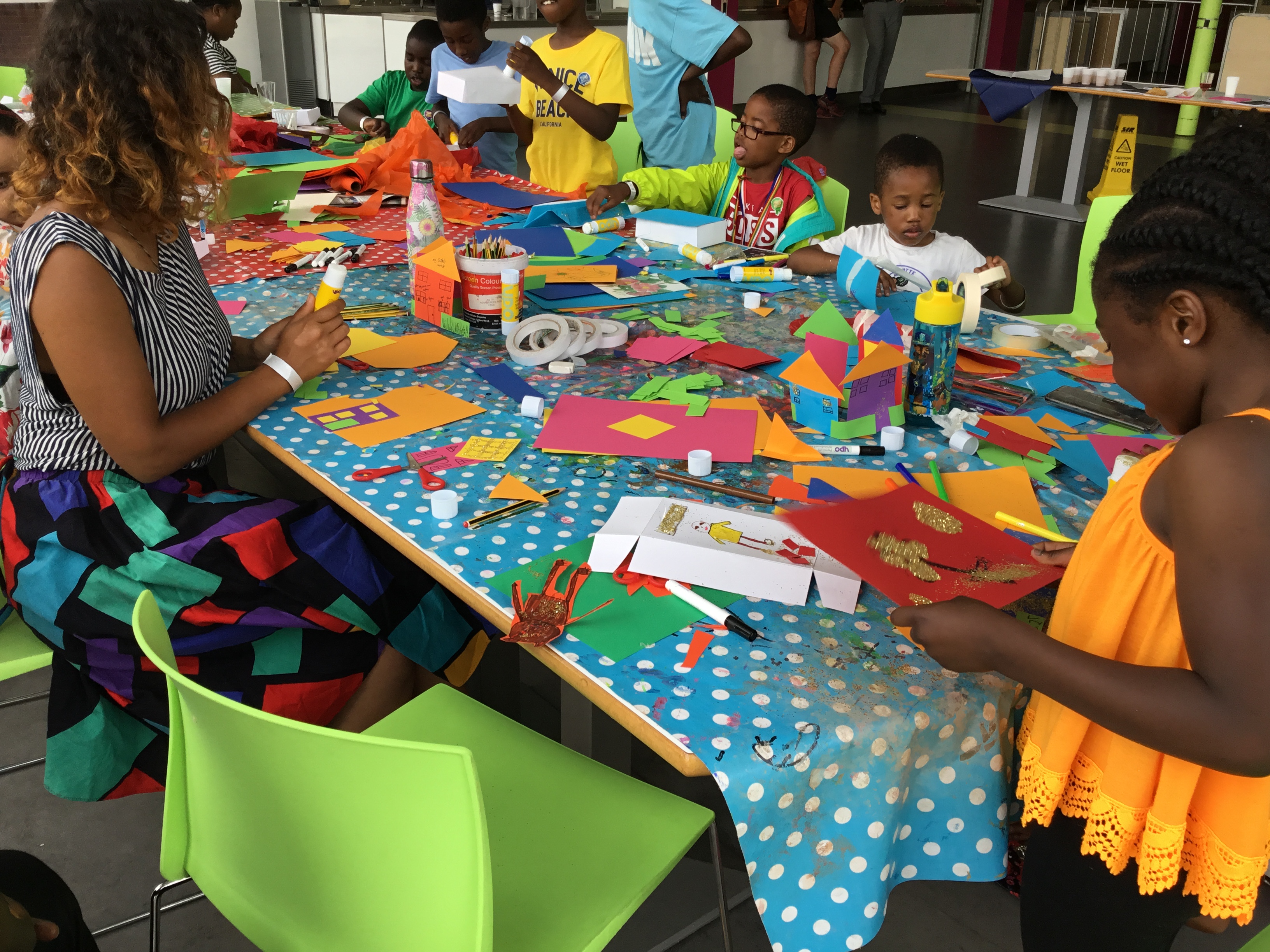 Photo of children and facilitator doing craft activities. There is lots of colourful card on a table covered with a blue and white polkadot tablecloth.