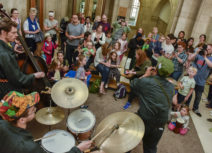 A man playing a clarinet, a man playing on a drum kit and a man playing a double bass perform in front of a group of children and families