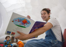 A woman with brown hair wearing denim dungarees and a white t-shirt is sitting reading a book to a group of children.