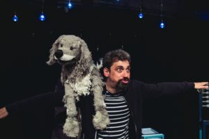 Production photo from Twinkle Twinkle. A grey Puppet dog sitting on the shoulder of an actor who is wearing a black and white striped top.