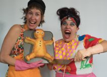 Two women smiling wearing colourful, mismatched clothing and holding a large gingerbread man style cake