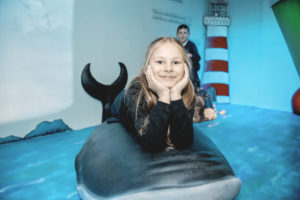 Young girl smiling and lying on a large plastic killer whale, in the background there is a young brown-haired boy and a painting of a lighthouse