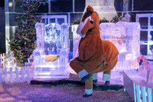 Picture of pantomime horse who is brown with a white nose and white lower legs, the horse is dancing in front of two thrones made of ice and a Christmas tree