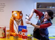 Photo of a woman with brown hair wearing a blue waistcoat and red long-sleeved t-shirt. She is in the middle of telling a story. She is stood next to a statue of The Tiger from Z-arts interactive exhibition. The Tiger is sat at a table, on the table is a box of Frosties and a carton of orange juice.