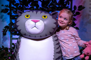Promo image for Meet Mog Live event. Photo of a young girl with blonde hair wearing a pink long-sleeved tshirt and denim shorts. She is standing next to a stature of Mog the Forgetful Cat which features in Z-arts interactive exhibition