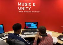 Lead image for songwriting competition. Two young people are sitting in front of a music keyboard and a computer screen, behind the screen is a red wall that says "music and unity" on it.
