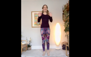 Lead image for Let's Yoga. Photo of a woman standing up wearing purple patterned leggings and a dark long-sleeved top waving at the camera.