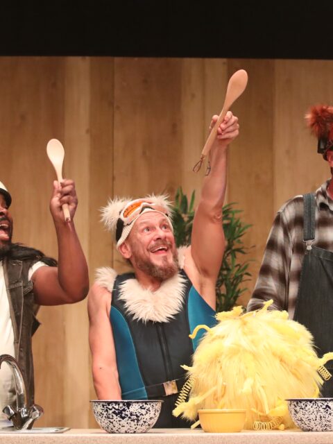 A stage scene from The Three Bears. Three performers are behind a kitchen cabinet. Panda, a bear inspired by a panda, is wearing black and white clothes with a black fur trim. Polar, a bear inspired by a polar bear, is wearing a blue ski suit, ski hat and goggles with white fur trim. Grizzly, a bear inspired by a grizzly bear, wears dungarees and a miner's hat with brown fur trim. Each bear holds up a wooden spoon On the kitchen cabinet are bowls and a large yellow ball of fluff (Goldie).