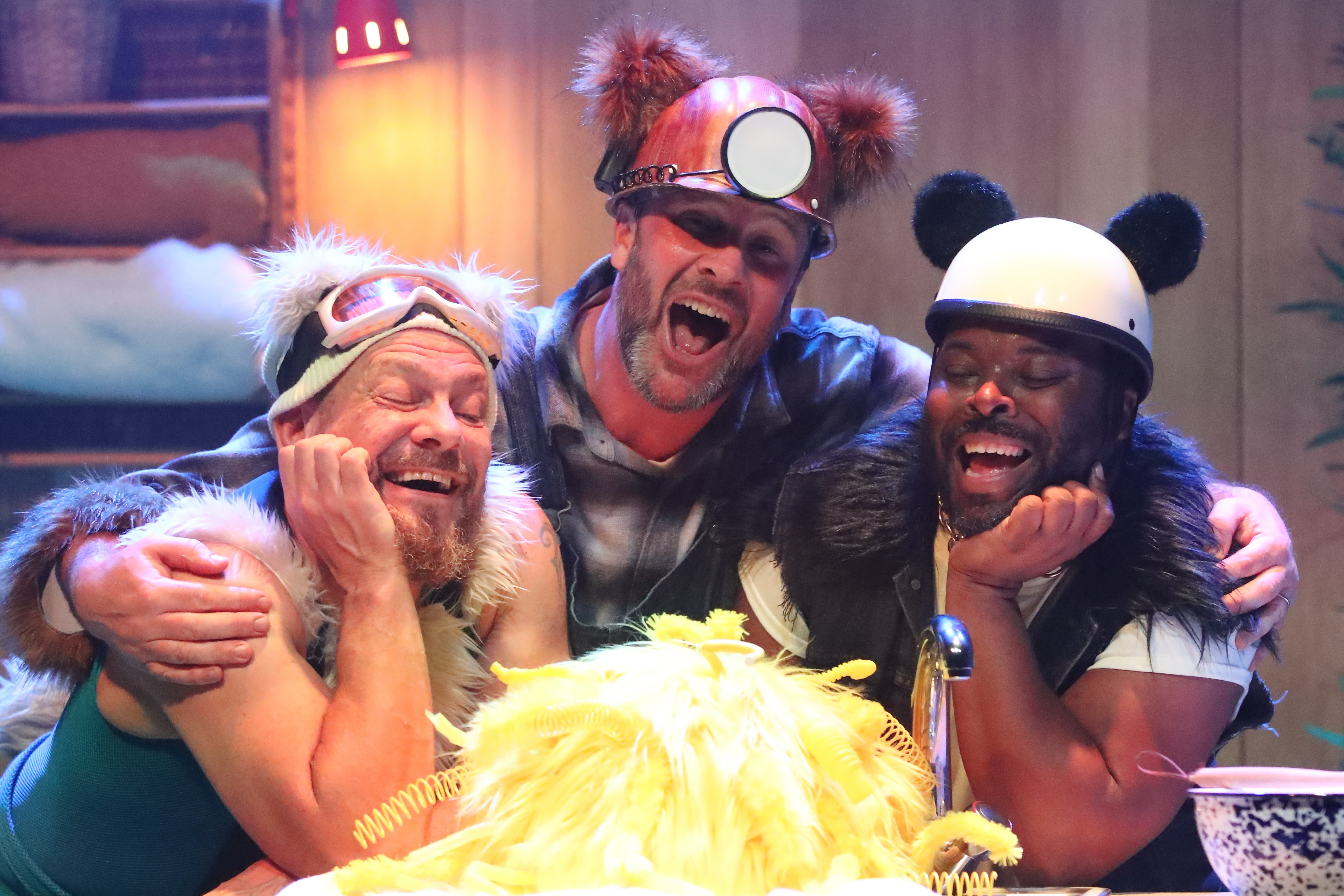 A stage scene from The Three Bears. Three performers are behind a kitchen cabinet. Panda, a bear inspired by a panda, is wearing black and white clothes with a black fur trim. Polar, a bear inspired by a polar bear, is wearing a blue ski suit, ski hat and goggles with white fur trim. Grizzly, a bear inspired by a grizzly bear, wears dungarees and a miner's hat with brown fur trim. They all look lovingly at ‘Goldie’ a mass of bright yellow feathers who is sat infant of them.